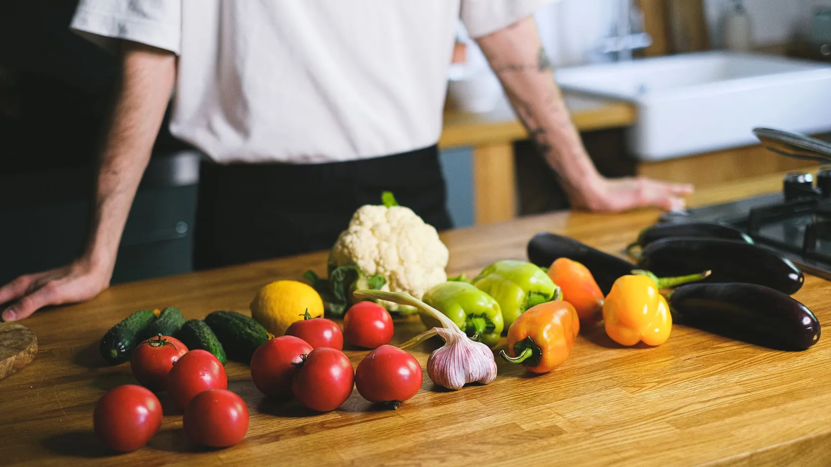 Tomates, concombres, poivrons, aubergines, chou-fleur, ail et citron posés sur un plan de travail en bois, devant une personne en tee-shirt blanc