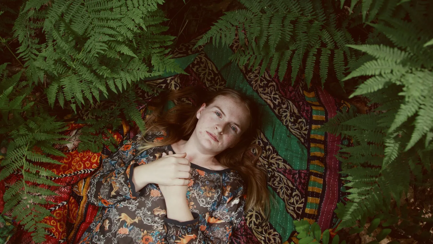 Femme allongée sur une couverture à motifs colorés, les mains croisées sur la poitrine, entourée de grandes fougères