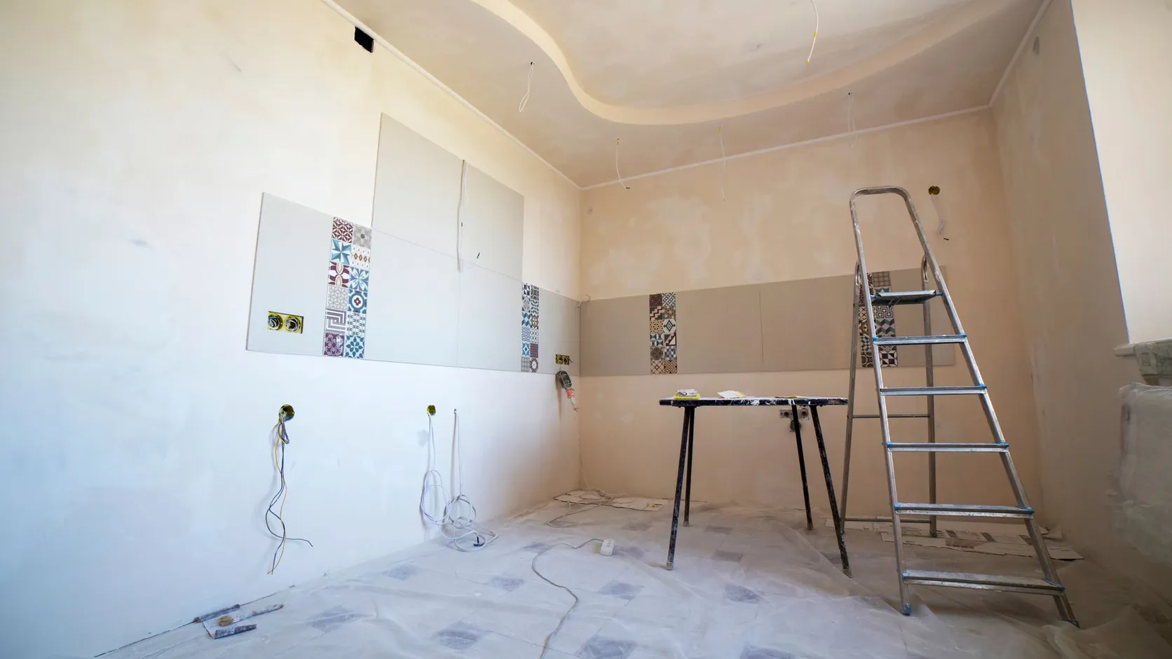 Pièce en rénovation aux murs enduits, avec frise de carreaux à motifs, fils électriques en attente, tréteau et escabeau sur un sol protégé