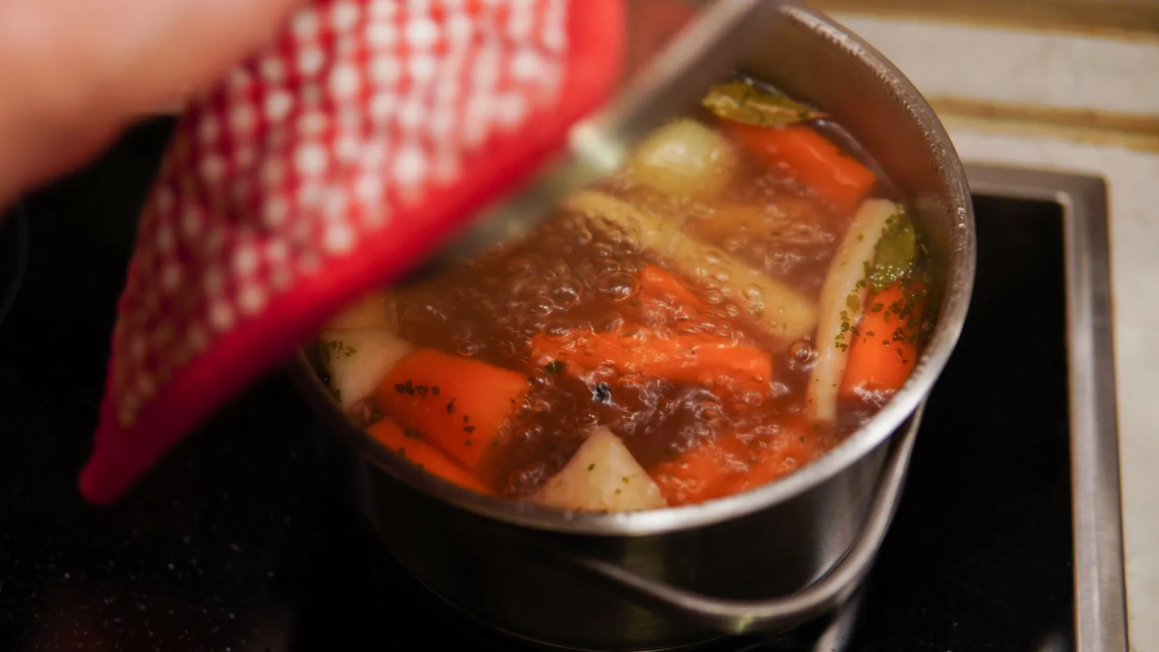 Marmite de bouillon frémissant avec carottes, navet et laurier sur une plaque de cuisson, une main gantée tenant le couvercle