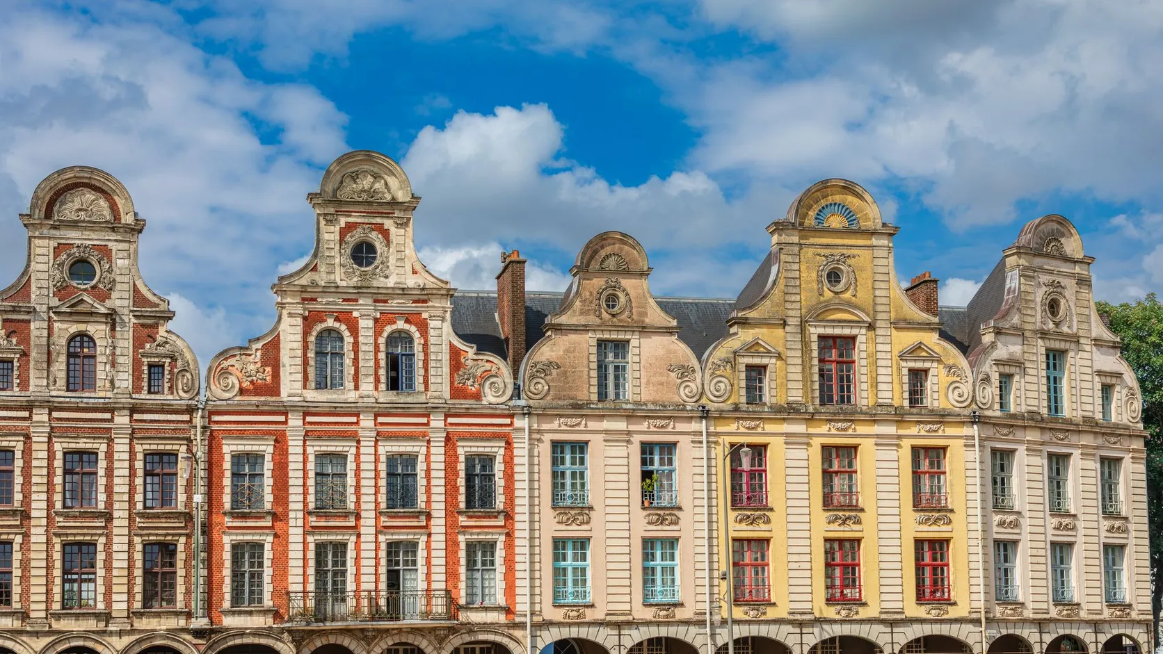 Façades à pignons baroques flamands, en brique rouge, ocre et pierre claire, sur une place d'Arras sous un ciel bleu nuageux
