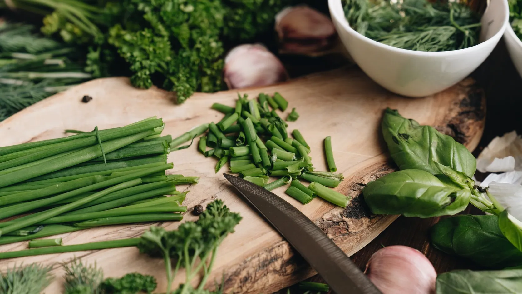 Ciboulette fraîchement ciselée sur une planche en bois, avec persil, aneth, basilic et gousses d'ail autour