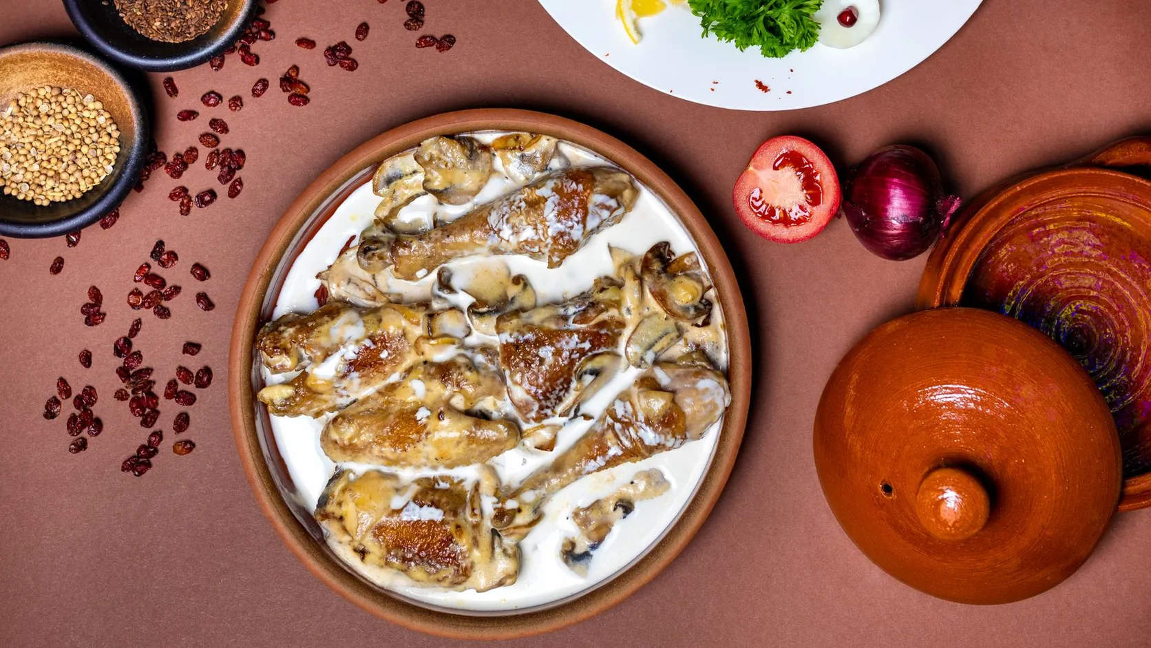 Morceaux de volaille dorés nappés de sauce blanche aux champignons dans un plat en terre, à côté d'une tomate, d'un oignon rouge et d'un couvercle de cocotte